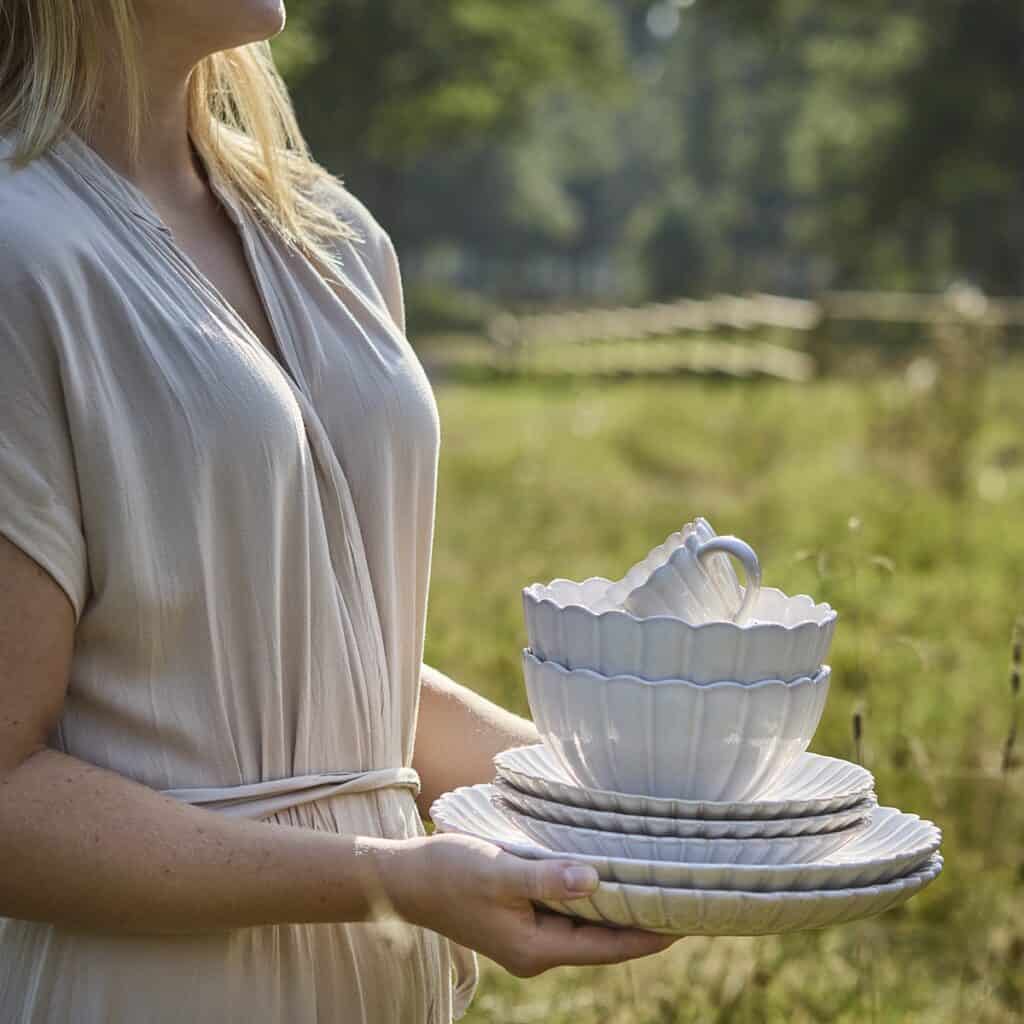En kvinna i ljus klänning håller en stapel av vackert vågiga skålar, fat och koppar i elfenbensvitt stengods från Affari of Sweden, omgiven av en lantlig sommaräng.