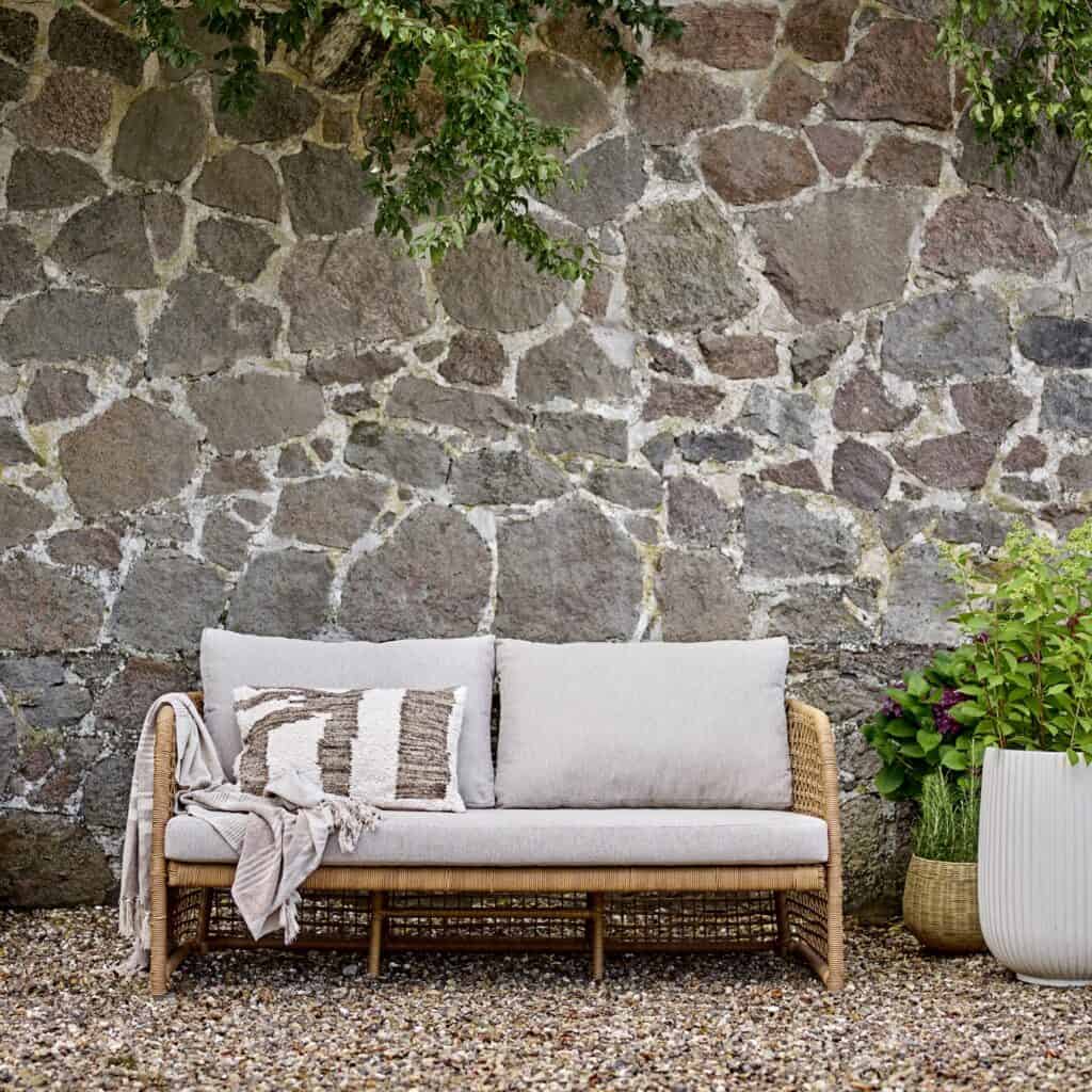Penzano soffa från Bloomingville i naturfärgad konstrotting med beige dynor, placerad mot en rustik stenmur och omgiven av grönska. En mysig och stilren utemöbel för trädgården eller uteplatsen.