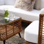 Närbild av Brafabs Covelo soffbord i naturfärgad konstrotting med glasbordsskiva, omgiven av en vitklädd soffa och fåtölj. Stilfullt och inbjudande för uteplatsen.