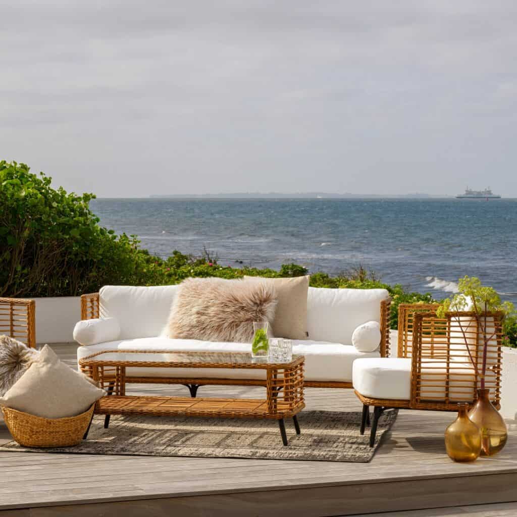 Ljus och inbjudande uteplats med Brafabs Covelo-serie i naturfärgad konstrotting. Soffbord, soffa och fåtölj med vita dynor skapar en stilren och avslappnad miljö vid havet.