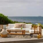 Ljus och inbjudande uteplats med Brafabs Covelo-serie i naturfärgad konstrotting. Soffbord, soffa och fåtölj med vita dynor skapar en stilren och avslappnad miljö vid havet.