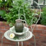 Ljusgrön trädgårdsstol COMO från Wikholm Form med dekorativa detaljer, använd som avlastningsyta med en grön keramikkanna och skål i ett växthus.