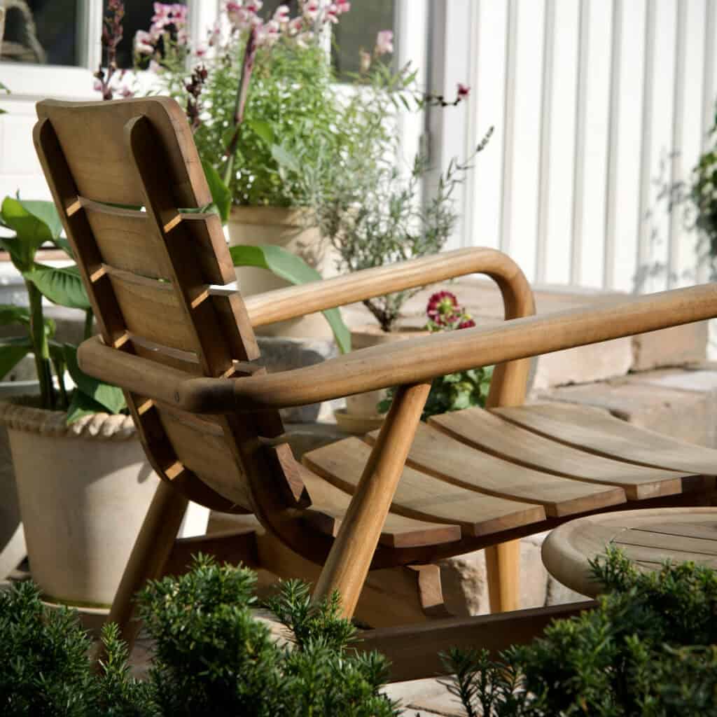 Loungefåtölj LILJA i teak placerad utomhus vid husfasad med gröna växter och krukor omkring.