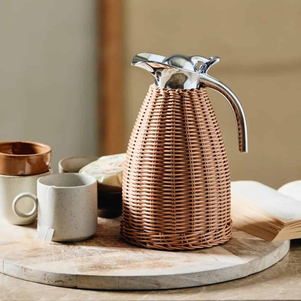 Termoskanna i rotting från Nordal placerad på ett rustikt träbord tillsammans med koppar och en bok.