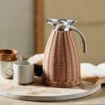 Termoskanna i rotting från Nordal placerad på ett rustikt träbord tillsammans med koppar och en bok.