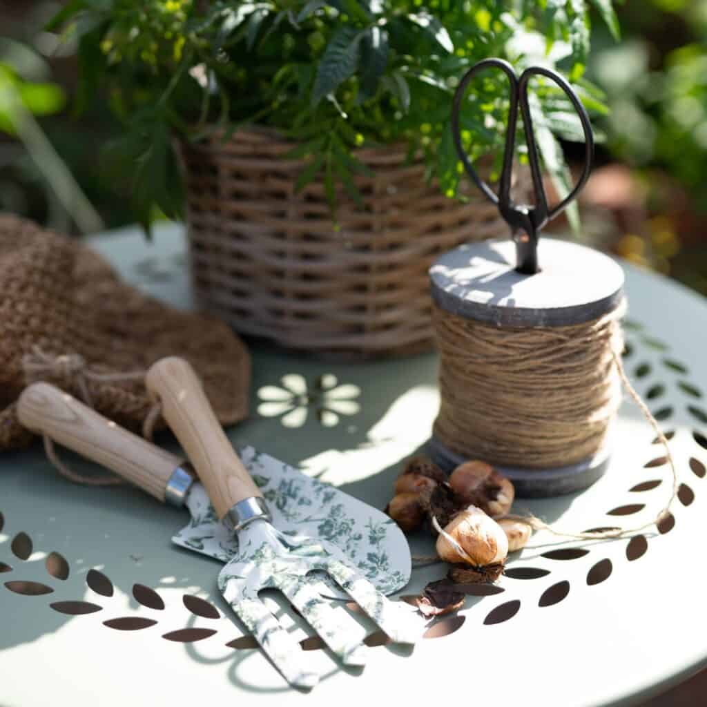 Närbild av ljusgrönt COMO bord från Wikholm Form, dekorerat med trädgårdsverktyg med blommigt mönster, jutesnöre, planteringslökar och en korgväxt i en solig trädgårdsmiljö.