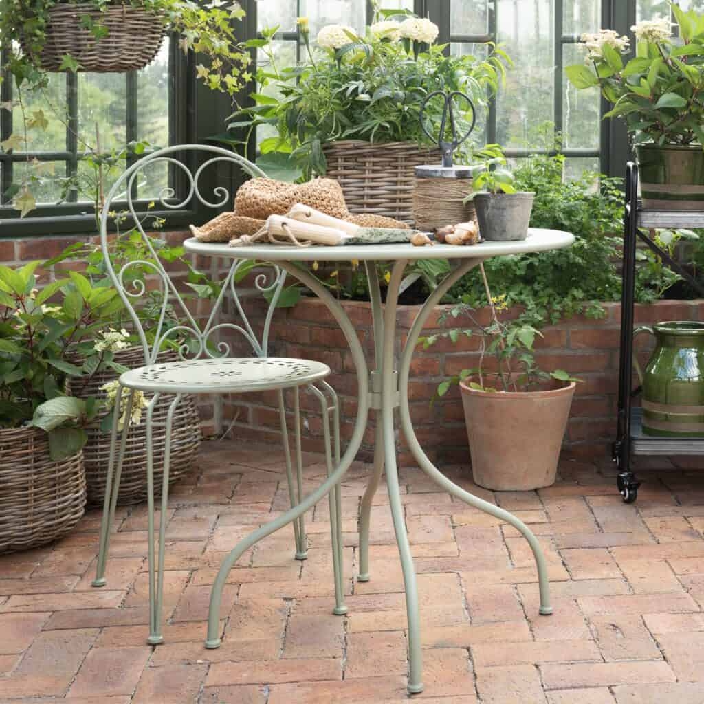 Ljusgrön trädgårdsstol och cafébord i smidesjärn från Wikholm Form, placerade i ett växthus med grönskande växter och terrakottakrukor.