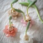 Fyra dekorativa glasblommor i olika färger används som hållare för spirande lökar, placerade på ett linnetyg för en naturlig och mjuk känsla.