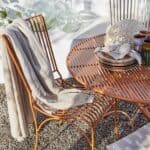 ELSA bordduk i beige bomull, elegant draperad över en stol vid ett utebord. Perfekt för avslappnade måltider i solen.