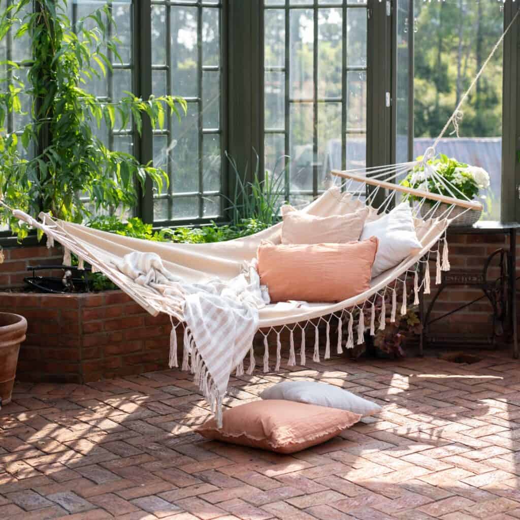 Hängmatta ARMIDA i offwhite upphängd i ett inglasat uterum, dekorerad med mjuka kuddar och en filt för en mysig och avkopplande atmosfär.