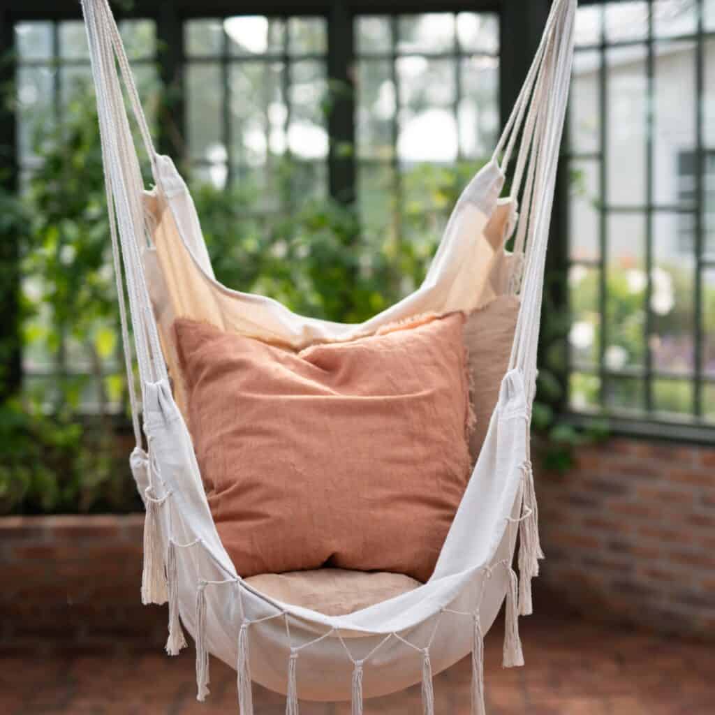 Hängstol ARMIDA i offwhite från Wikholm Form, upphängd i ett mysigt växthus med gröna växter i bakgrunden. Stolen är dekorerad med jordnära kuddar för extra komfort.