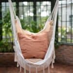 Hängstol ARMIDA i offwhite från Wikholm Form, upphängd i ett mysigt växthus med gröna växter i bakgrunden. Stolen är dekorerad med jordnära kuddar för extra komfort.