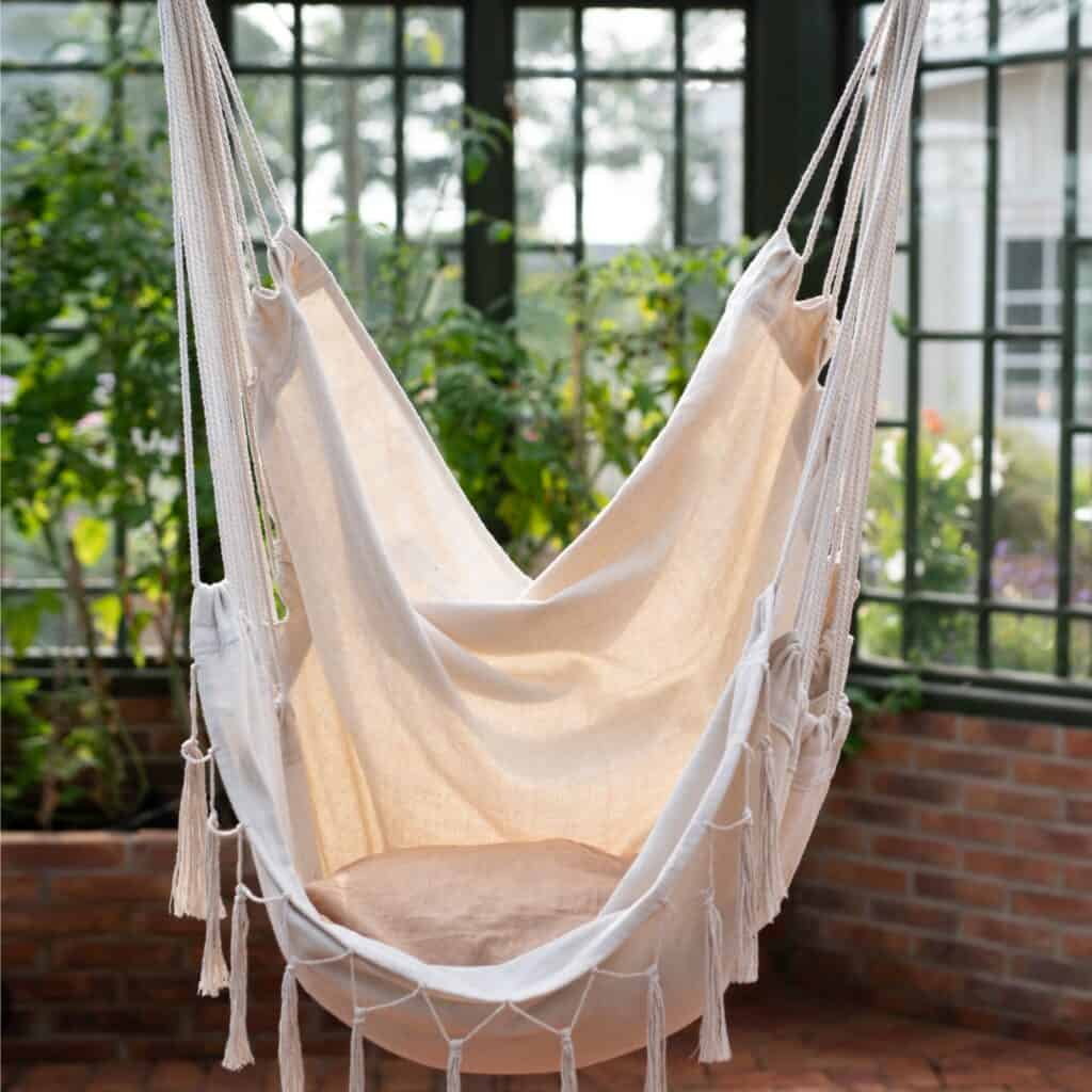 Hängstol ARMIDA i offwhite från Wikholm Form, upphängd i ett ljust och grönskande växthus. En stilren och bohemisk sittplats för avkoppling.