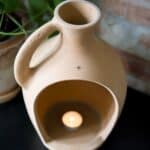 Närbild av Floda Lykta i terrakotta från Affari of Sweden, sedd uppifrån med ett värmeljus inuti, mot en bakgrund av tegelvägg och gröna växter.