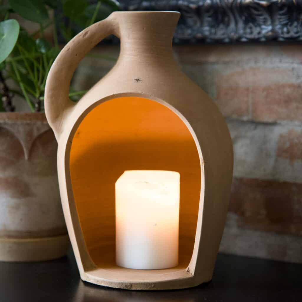 Floda Lykta i terrakotta från Affari of Sweden med ett tänt blockljus inuti, sprider ett varmt och stämningsfullt sken i en rustik miljö.