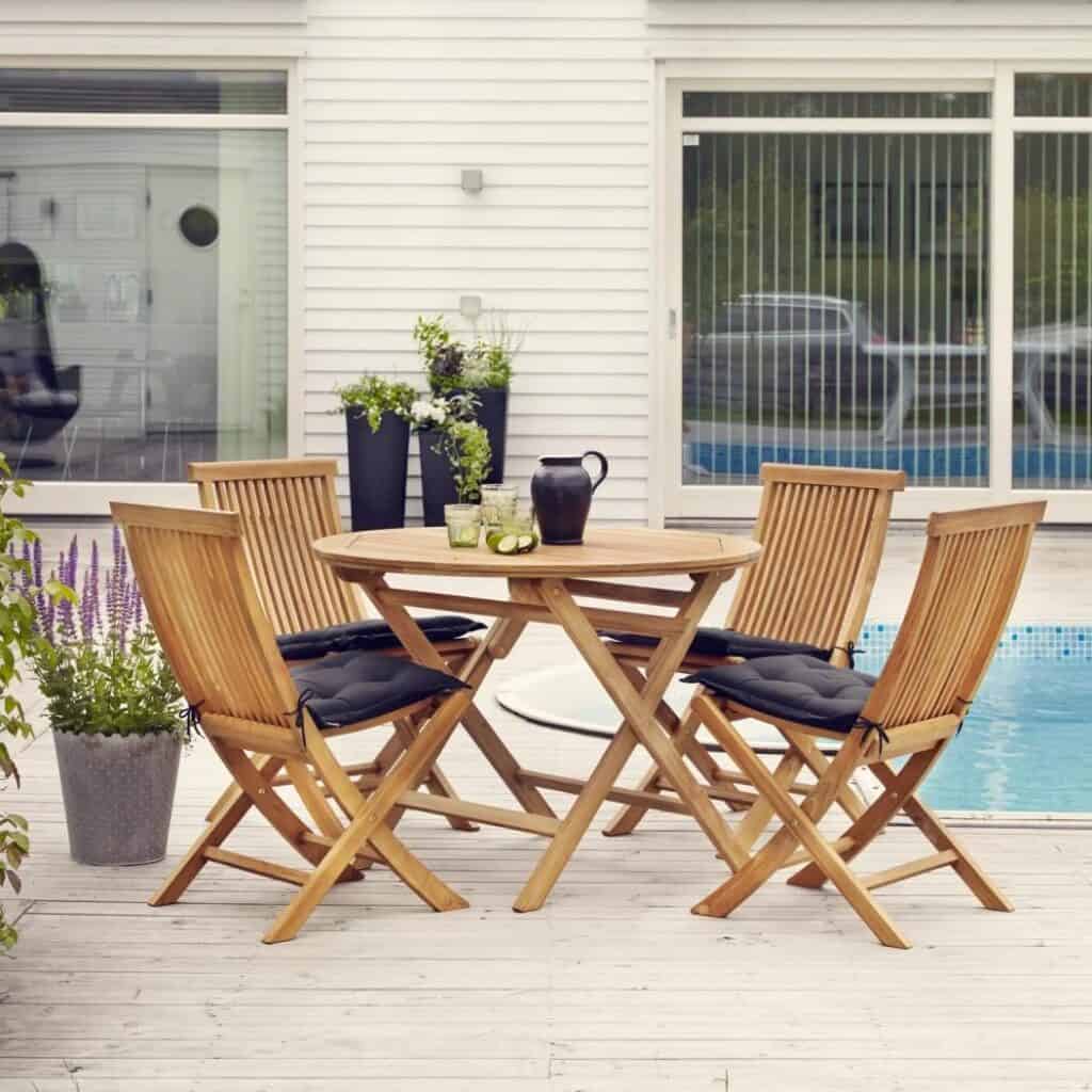 Runt matbord TURIN 100 cm i teak från Brafab, omgiven av fyra matchande teakstolar med svarta dynor, placerat på en altan vid en pool.