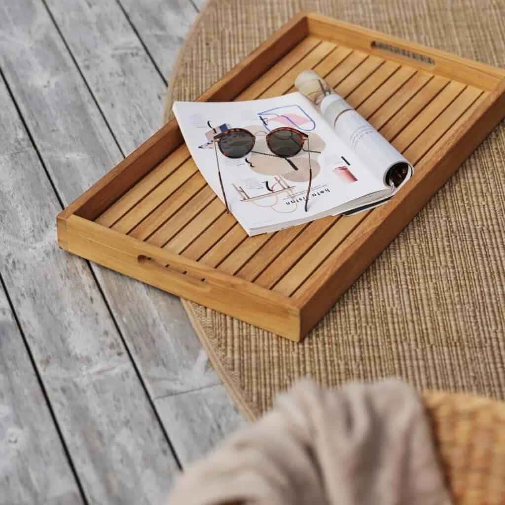 Serveringsbricka TURIN i teak från Brafab placerad på ett bord utomhus, med en tidning och solglasögon ovanpå. En stilren och praktisk detalj för servering och dekoration.