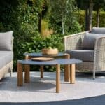 Två Brafab Domino soffbord i antracit och teak, stilfullt placerade på en uteplats med rottingmöbler och grönskande omgivning.