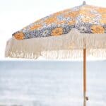 Parasoll Gatsby blommig 185 cm från Brafab med fransar och retrotyg i gult, blått och vitt uppställt vid havet.