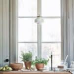 Taklampa med tvinnad sladd i mässing och vit opalglas från Strömshaga, upphängd framför ett fönster i lantligt kök.