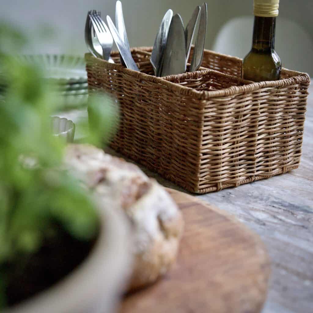 Förvaringskorg i rotting med fack från Madam Stoltz, fylld med bestick och flaska olivolja, stående på ett marmorbord.
