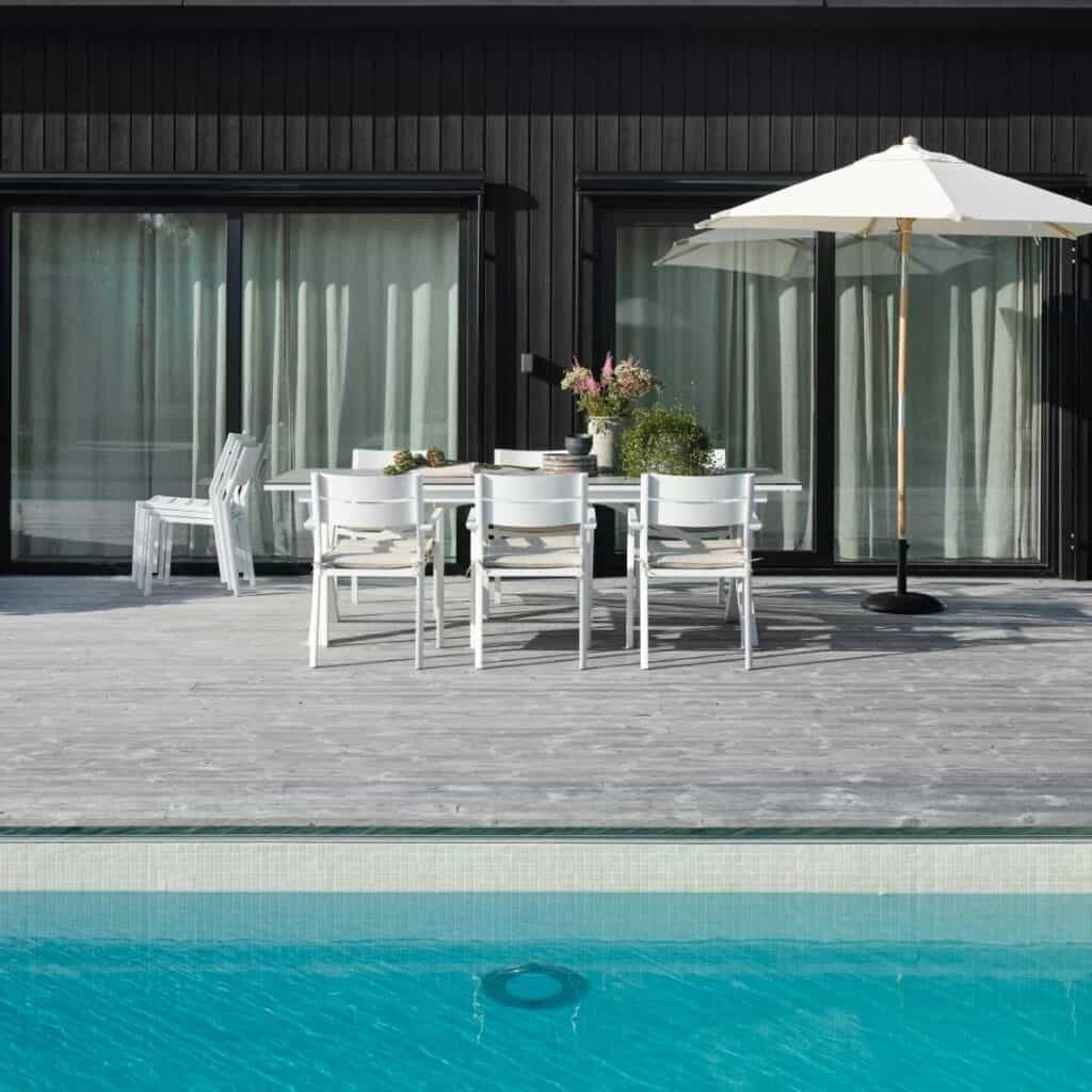 Matplats vid pool med vita Delia karmstolar från Brafab, utomhusbord och parasoll på grå altan framför svart husfasad.