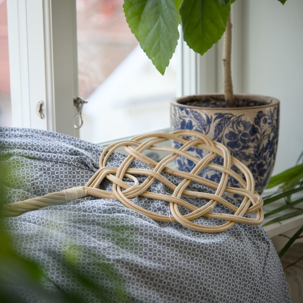 Mattpiska i rotting på tyg vid fönster med växt i bakgrunden