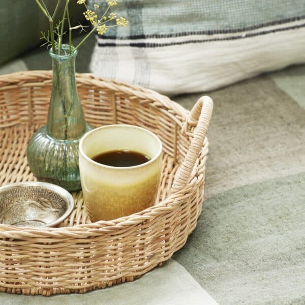 Rund bricka i rotting från Madam Stoltz med kopp kaffe, liten skål och glasvas med blomma på ett dukat bord.