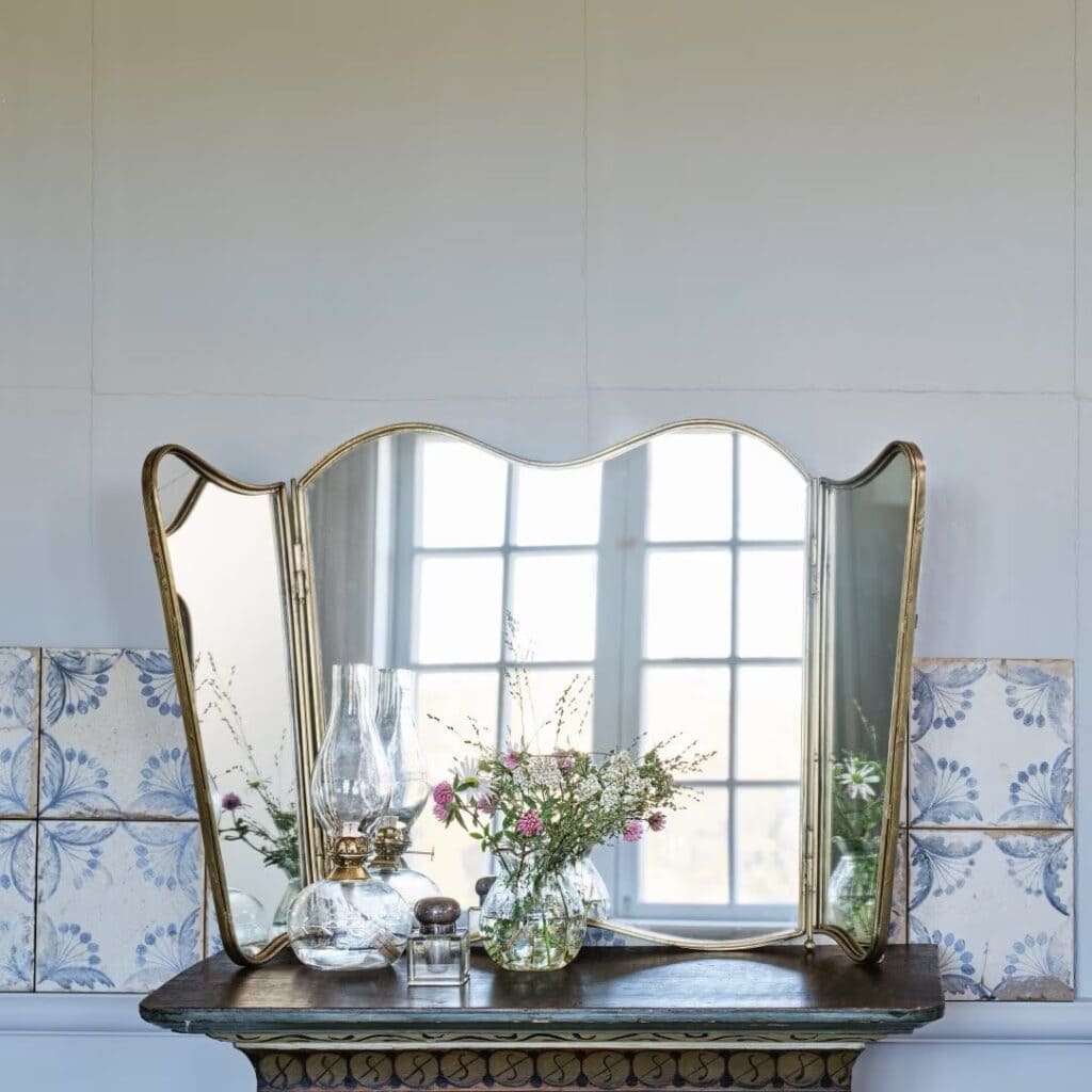 Vikbar spegel i antik mässing från Strömshaga, placerad på ett gammaldags bord med dekorativa blommor och oljelampa framför. Spegeln reflekterar ett fönster som skapar ett ljust och luftigt intryck.