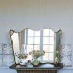 Vikbar spegel i antik mässing från Strömshaga, placerad på ett gammaldags bord med dekorativa blommor och oljelampa framför. Spegeln reflekterar ett fönster som skapar ett ljust och luftigt intryck.