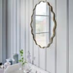 Oval spegel med vågig kant i antik mässing från Strömshaga, upphängd på pärlspontpanel i badrum med handfat och ljusinsläpp.