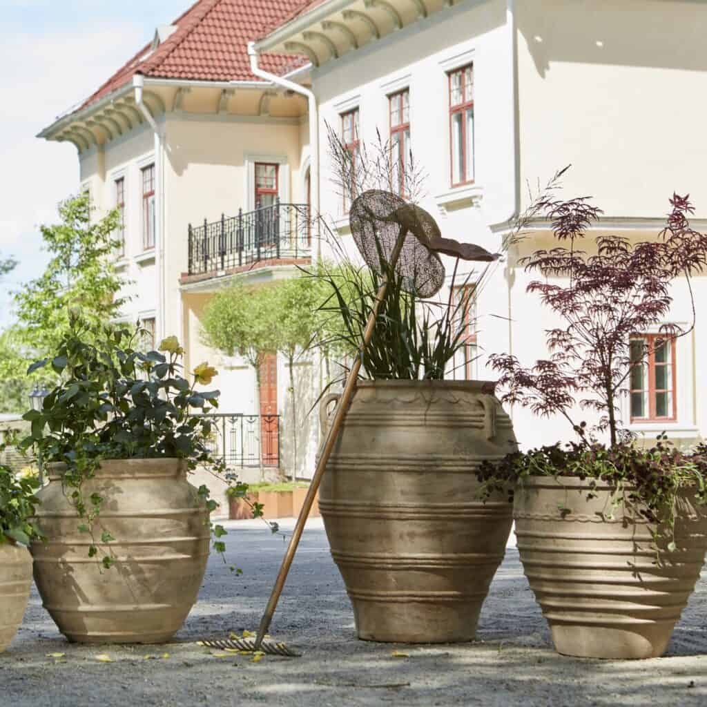 Vackra urnor i antik terrakotta från Wikholm Form, inklusive modellen TALIA. Perfekt för att skapa en klassisk och tidlös atmosfär i trädgården eller på uteplatsen. Planterade med gröna växter och omgivna av en harmonisk miljö vid en elegant villa.