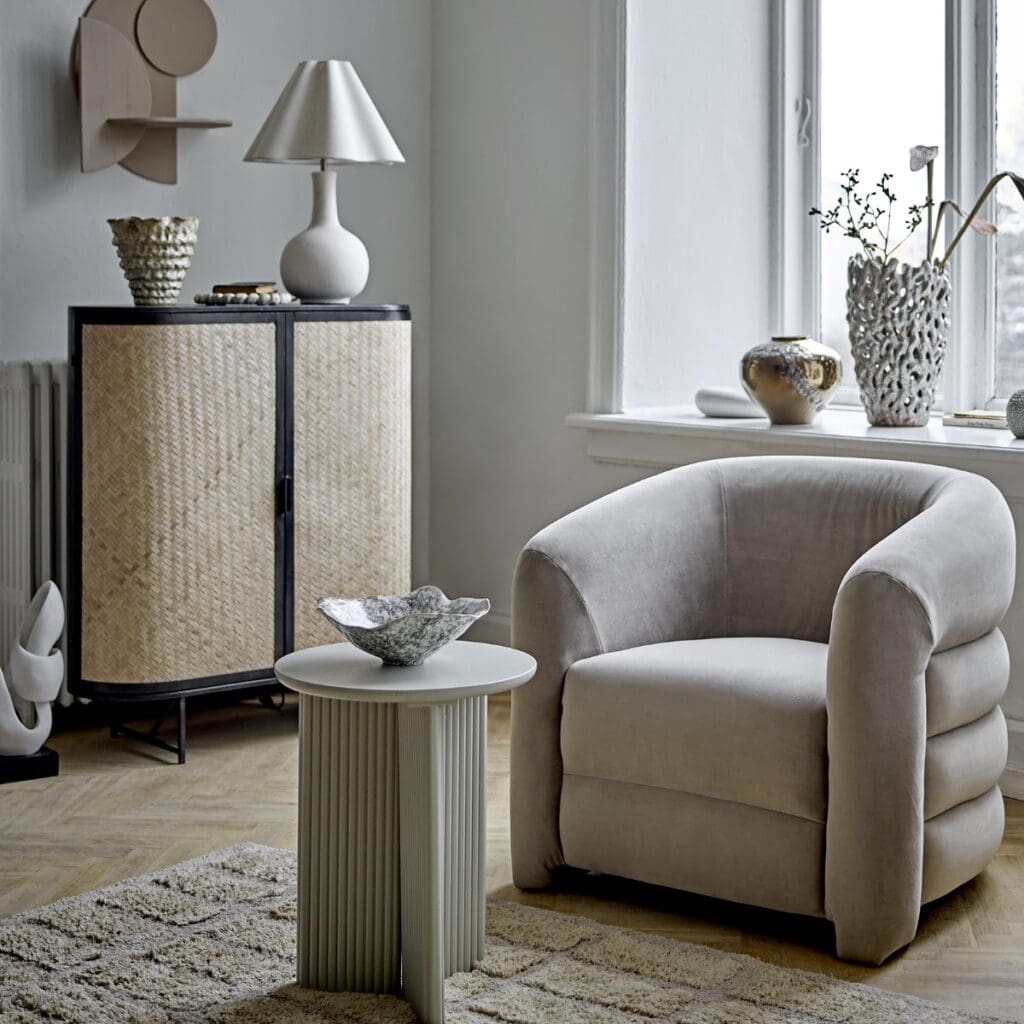 Skandinaviskt vardagsrum med skänk i svart metall och rotting från Bloomingville, kombinerat med fåtölj i beige sammet och ljusa inredningsdetaljer. Finns hos Hemlängtan.