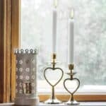 Värmeljuslykta kakelugn vit från Strömshaga på fönsterbräda, bredvid två mässingsljusstakar med hjärtform.