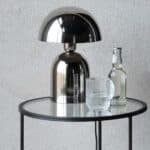 Bordslampa i krom – ROME från Wikholm Form på glasbord, i modern miljö med dryck och glas bredvid.