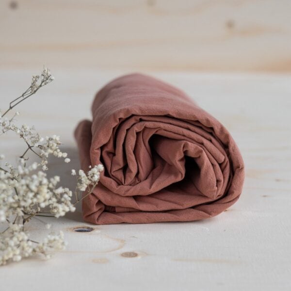 Underlakan ekologisk bomull Terracotta 240×260 cm – hoprullat på träbord med vita blommor i förgrunden