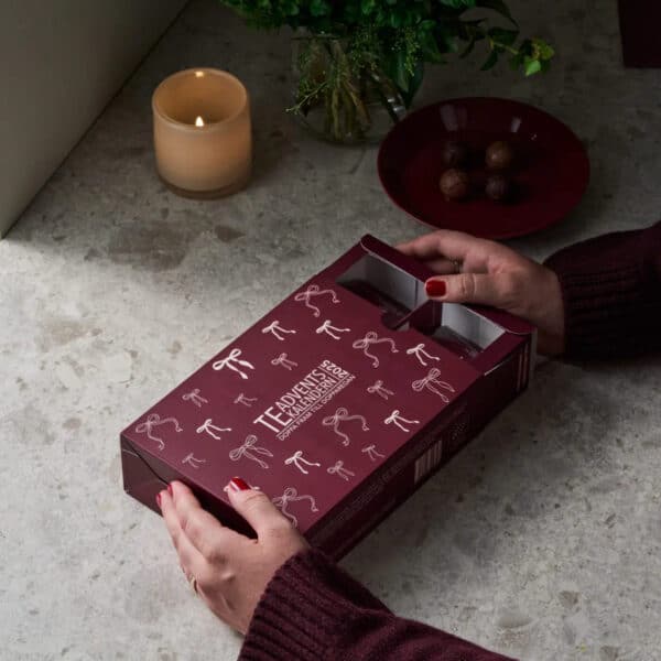 Person öppnar NABOs Teadventskalender 2025 – vinröd ask med pudderrosa rosetter, stående på ett bord med tända ljus och chokladpraliner.