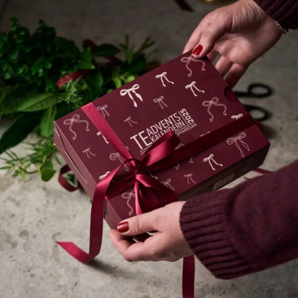 Person knyter ett vinrött sidenband runt NABOs Teadventskalender 2025 – en vinröd ask med pudderrosa rosetter, perfekt som julgåva.