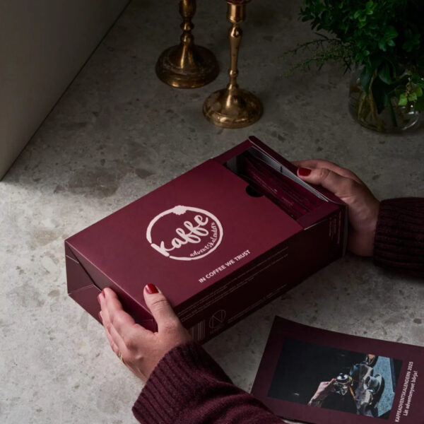 Händer som öppnar Nabos vinröda kaffeadventskalender med präglad logga – 24 kaffesorter i elegant ask på stenskiva.
