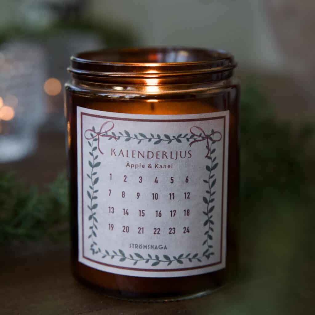 Kalenderljus med doft av äpple och kanel i mörkrött glas, tänt och placerat på ett bord med gröna kvistar i bakgrunden.