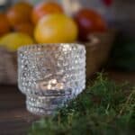 Glaslykta i strukturerat glas med tänt ljus, placerad på ett träbord med granris och citrusfrukter i bakgrunden.