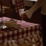 Närbild av juldukning med rödvit rutig duk i ekologisk bomull, tända ljus och hand som lyfter vinglas – Duk Ida Ruta Röd/Vit från Strömshaga.