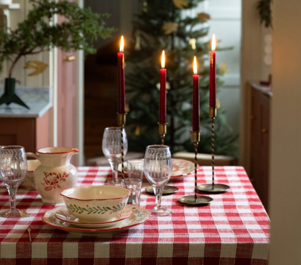 Ljus i mässingsljusstakar på rödvitrutig duk – lantlig juldukning med porslin och gran i bakgrunden.