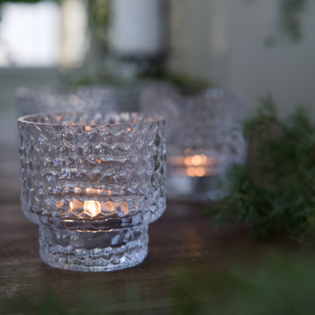 Ljuslykta DOTTI i klart bubbligt glas med tänt värmeljus, placerad på träbord med gröna kvistar i bakgrunden.