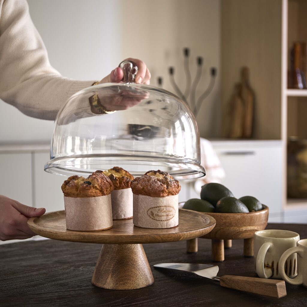 Kakfat i mangoträ med glaskupa BELLA, muffins och varma trädetaljer på bord.