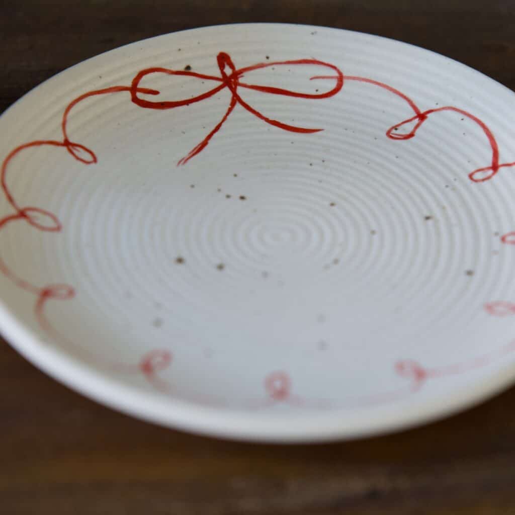Närbild av assiett i vit keramik med handmålad röd rosett runt kanten.