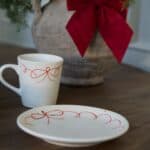 Kopp och assiett i vit keramik med handmålade röda rosetter, stylade på träbord framför julrosett.