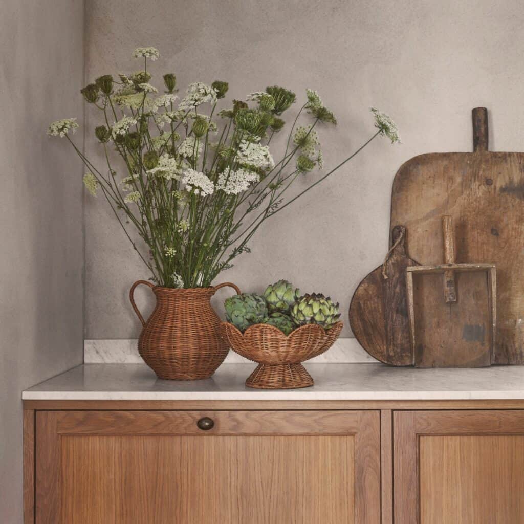 Fruktskål i naturfärgad rotting på fot placerad på köksbänk tillsammans med rottingkärl och trädetaljer