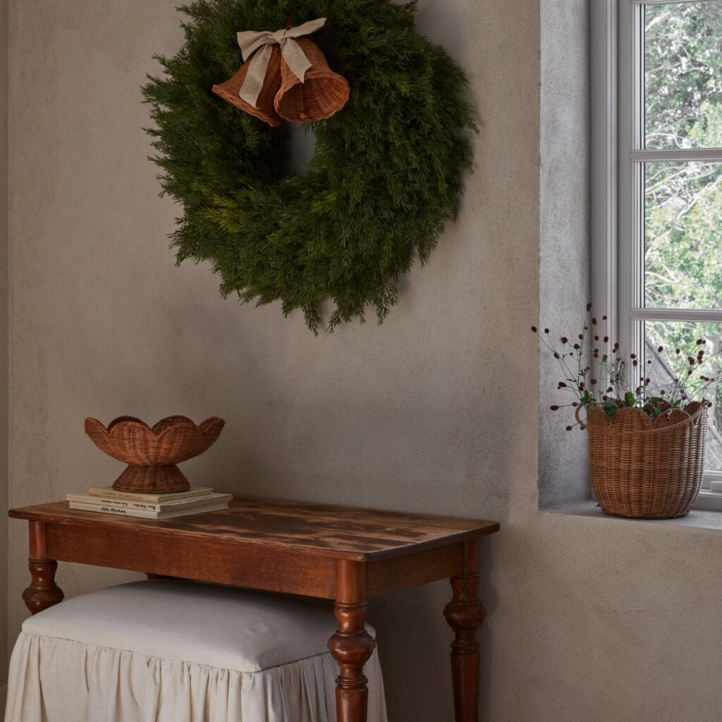 Fruktskål i naturfärgad rotting på fot placerad på litet bord i varm miljö med grön krans på väggen