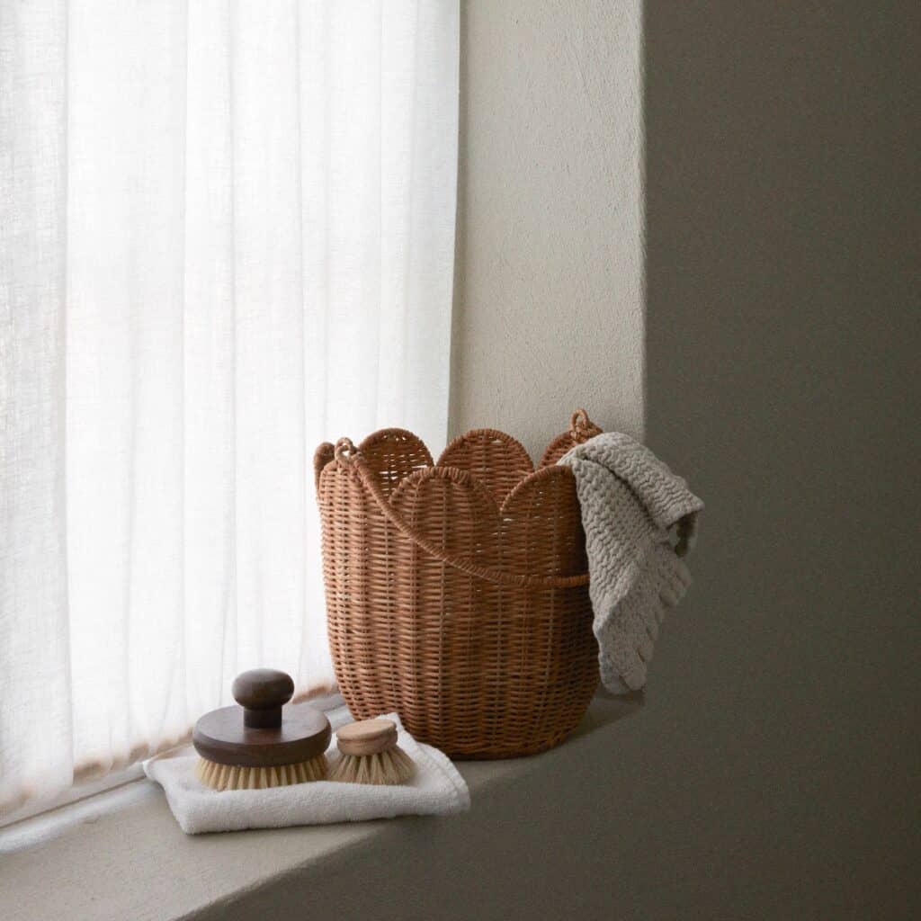 Handgjord korg i naturfärgad rotting med vågig kant placerad på fönsterbräda tillsammans med borstar och textilier.